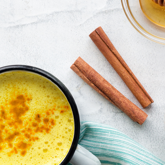 A spiced golden cacao latte in a mug next to cinnamon two sticks, honey and a bowl of cacao powder on a marble kitchen surface.