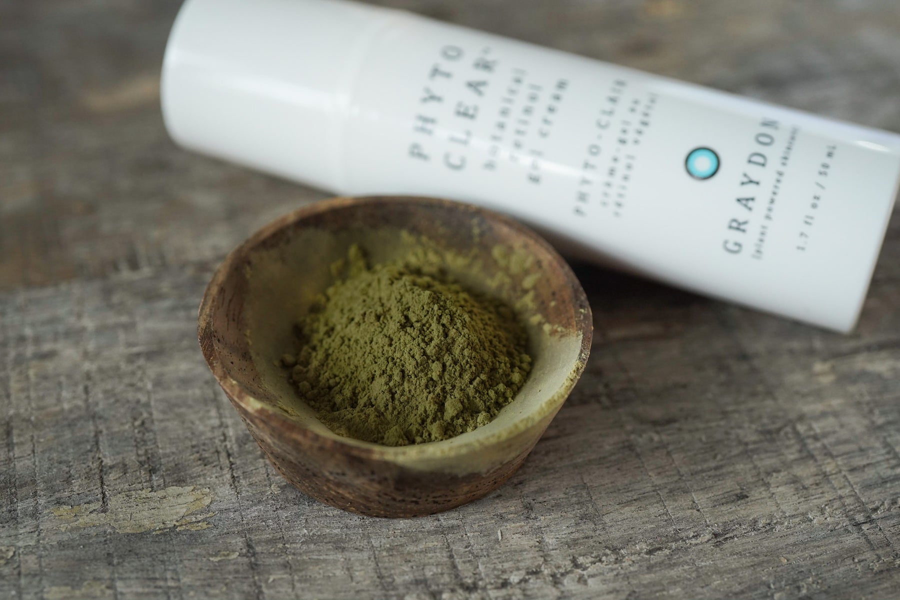 A natural face moisturizer containing chlorella in a white bottle next to a small brown wooden bowl of powdered chlorella sitting on a rustic surface. 