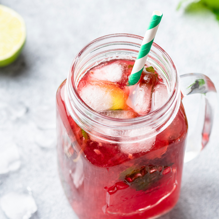 Mason jar mug with ice and bright red liquid on a white counter top with peppermint sprigs, lemon wedges and ice cubes in the background.