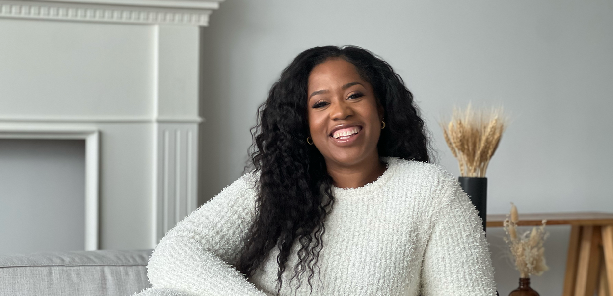 A smiling Jhanelle Peters sitting sideways on a couch, holding a camera