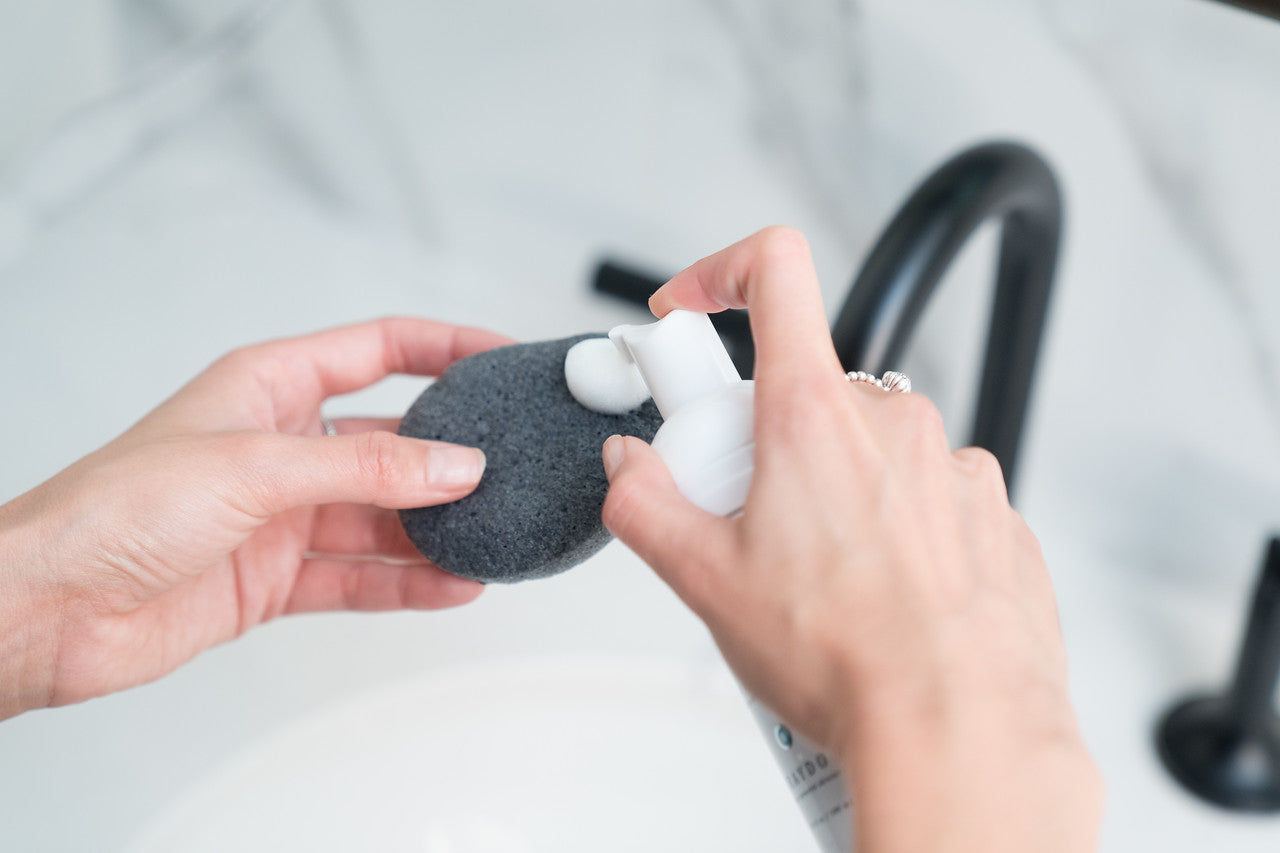 Close-up of a woman dispensing a white foaming cleanser for combination skin onto a black-coloured bamboo charcoal sponge for sensitive skin with a modern-looking sink in the background.