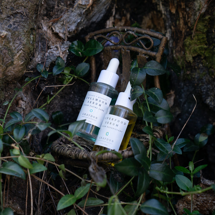 Two serums in glass bottles placed on wooden platform covered with leaves.