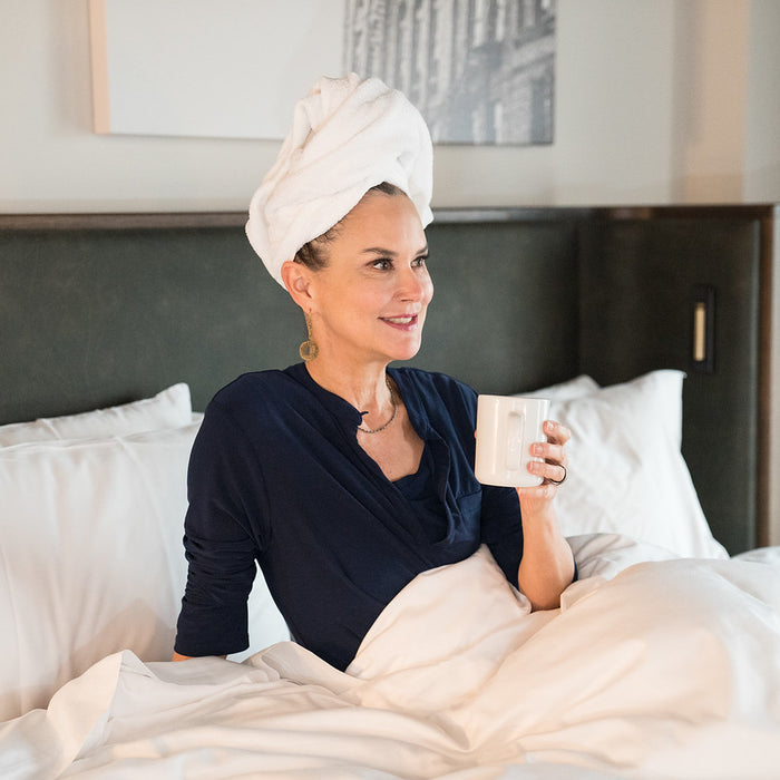 A woman sitting in a hotel bed holding a cup of coffee in her hand with her hair wrapped in a towel