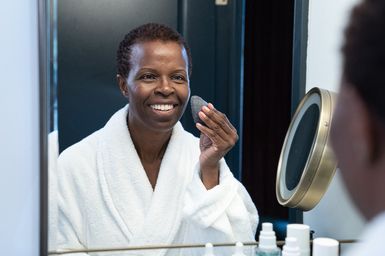 A dark-skinned woman exfoliating her skin with a konjac sponge for sensitive skin in front of a mirror