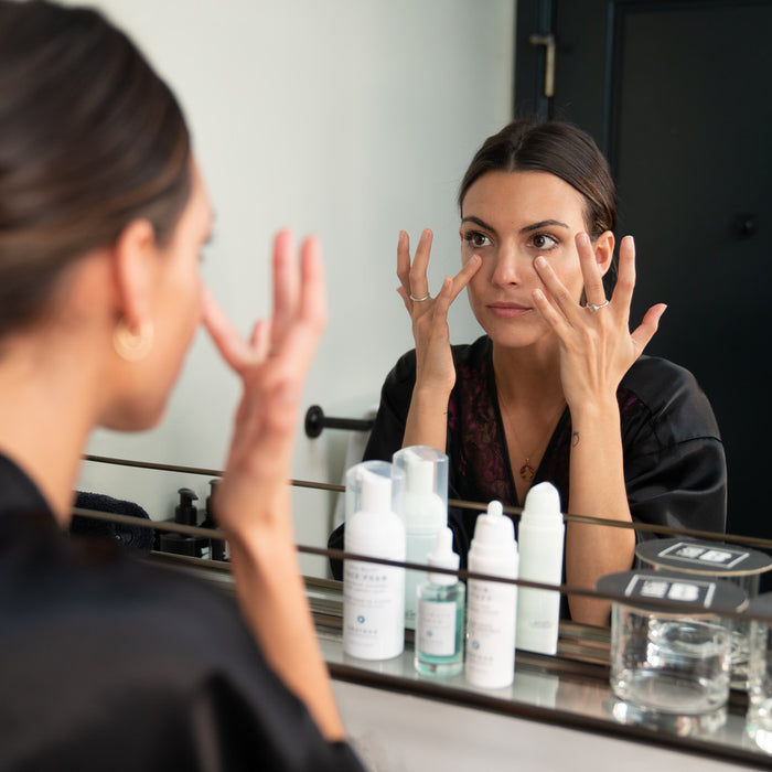 A close-up of woman with her hair tied back in a ponytail applying skincare to her face in front of a mirror using both hands.