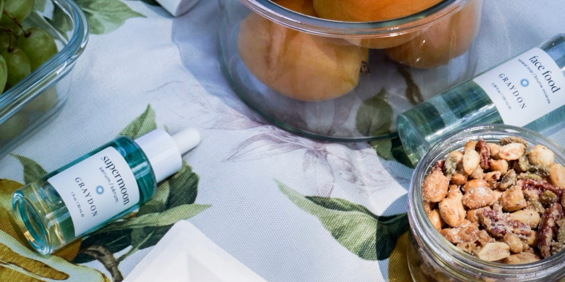 skincare serum and face mist on picnic blanket alongside some summer snacks