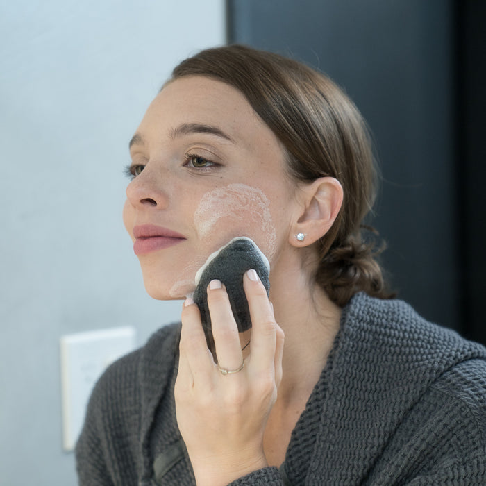 A woman cleansing her skin using konjac sponge for sensitive skin in front of a mirror.