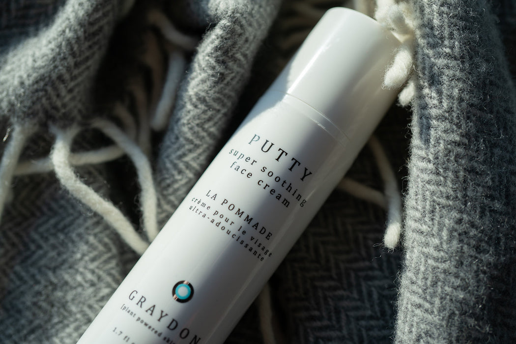 A bottle of moisturizer for dry, irritated skin lying on a grey and white herringbone blanket. 