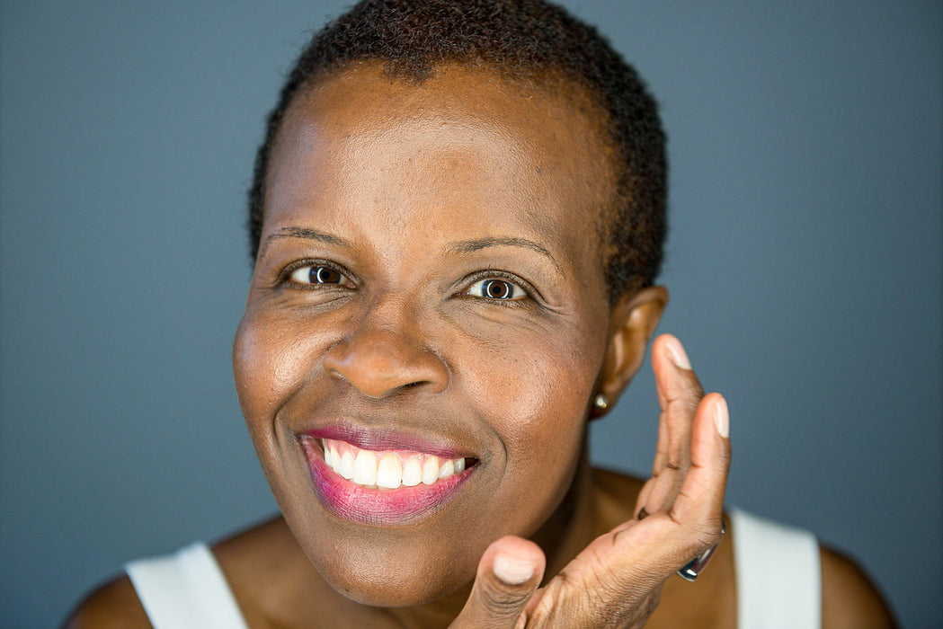 A smiling dark-skinned woman applying a tinted moisturizing skin primer with a blue-grey wall in the background. 