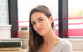 A smiling olive-skinned woman with long brown hair and glowing skin with books and a cactus plant in the background. 